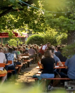 Besucher genießen den Sixengarten auf der Nördlinger Mess unter schattigen Bäumen. In der Abendsonne sitzen Gäste an langen Biergartentischen und verbringen gemeinsame Stunden bei Bier und regionalen Schmankerln.