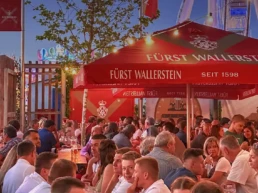 Voller Sixengarten auf der Nördlinger Mess bei Abendstimmung: Besucher sitzen unter den roten Fürst-Wallerstein-Schirmen, genießen Bier und regionale Schmankerl, während im Hintergrund die Lichter der Fahrgeschäfte leuchten.