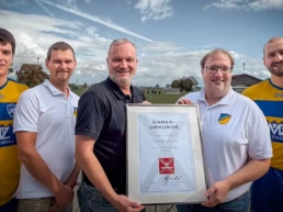 Harald Schmidt vom Fürst Wallerstein Brauhaus überreicht Vertretern des FC Birkhausen eine Ehrenurkunde für 50 Jahre Partnerschaft. Zwei Fußballspieler in gelb-blauen Trikots begleiten die Ehrung am Sportplatz.